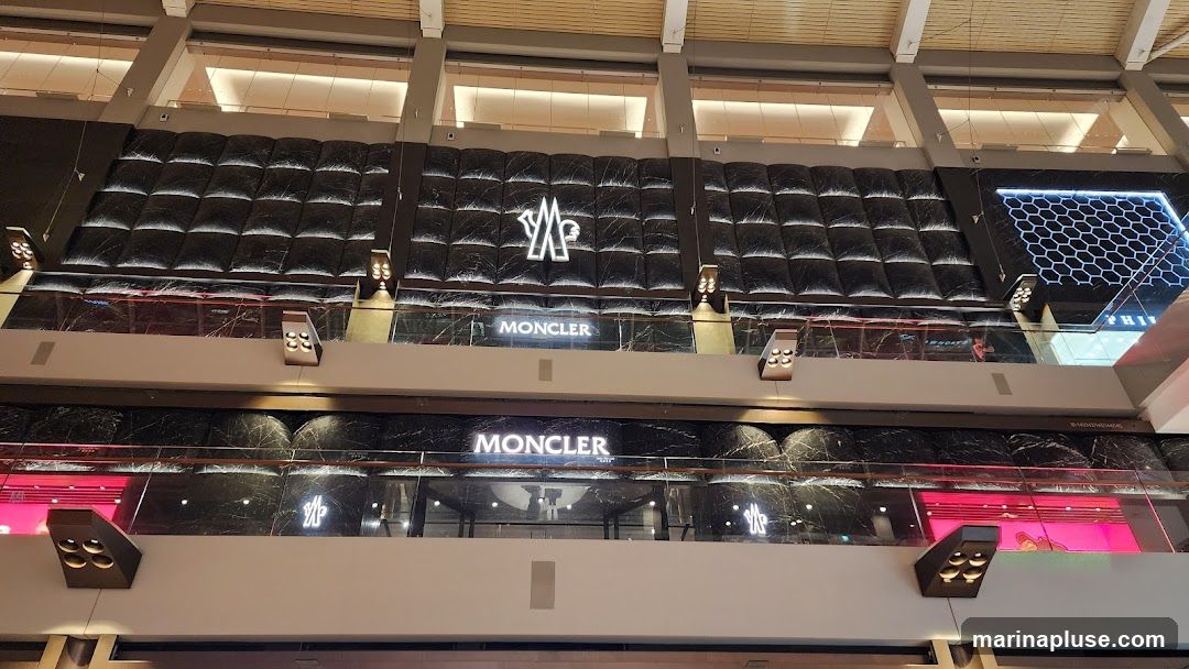 A wide shot of a Moncler store facade on two levels of a shopping mall. The upper level features a large Moncler logo and the word "MONCLER" in illuminated signage. The lower level also displays "MONCLER" in illuminated signage, with a glimpse of the store interior visible through the glass. The facade is decorated with dark, textured panels and marble accents.