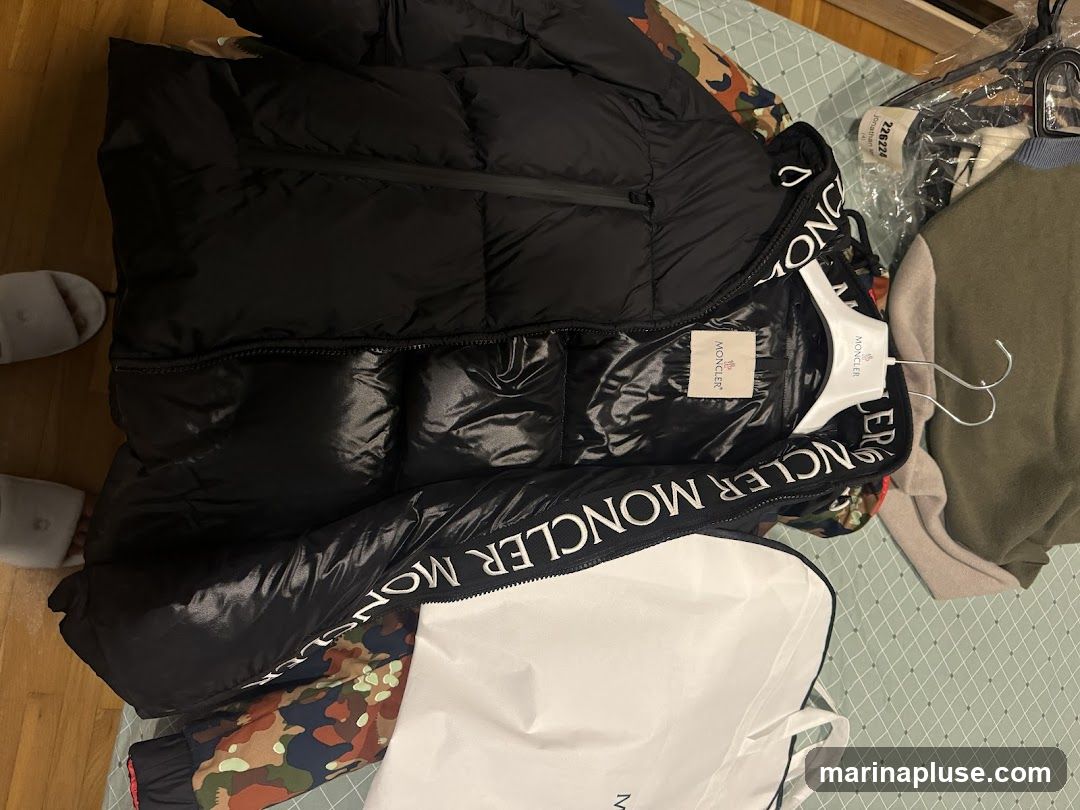 A black Moncler puffer jacket is laid out on a surface, with its hood open. The jacket features a zipper closure and a prominent Moncler logo tag inside. The words "MONCLER MONCLER" are printed repeatedly along the edge of the hood. Other clothing items, including a green sweater and a camouflage-patterned garment, are visible in the background, along with a white garment bag.