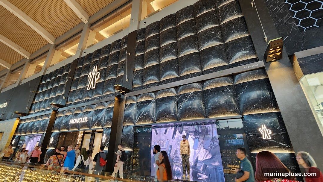 The exterior of a Moncler store with a distinctive black marble-patterned facade. The store features large illuminated Moncler logos and signage above the entrance. People are visible walking in front of the store, and a digital display shows a mannequin.