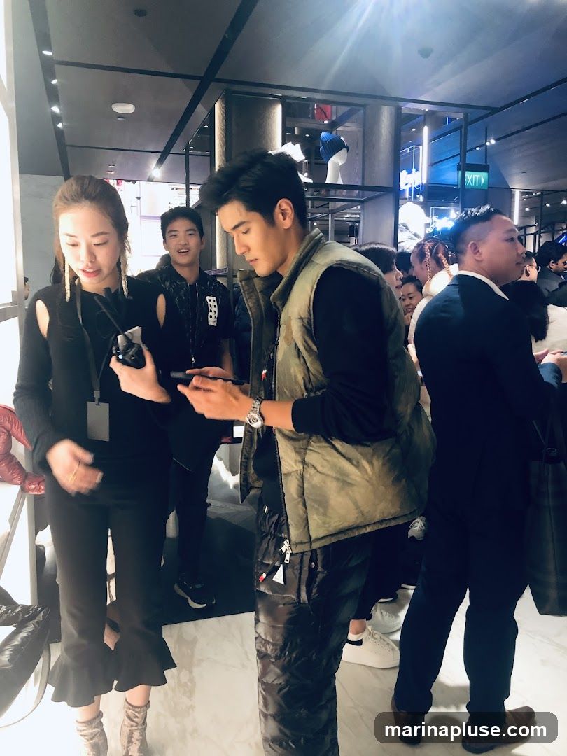 A group of people are gathered inside a store, with some looking at their phones. A woman in black pants with ruffled hems is holding a device, and a man in a green vest and puffer pants is looking at his phone. In the background, there are shelves displaying clothing and accessories, and a sign with an arrow pointing to the right that says "EXIT".