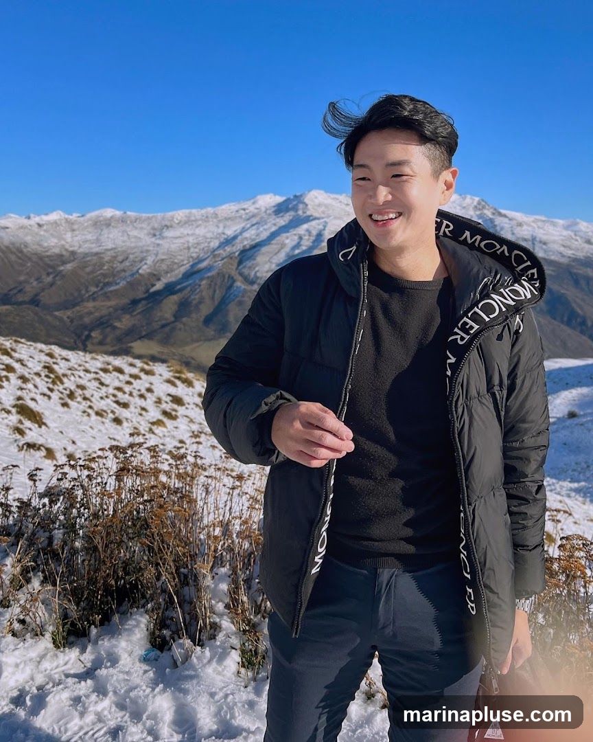 A young man smiles while standing in a snowy mountainous landscape. He is wearing a black puffer jacket with white lettering that reads "MONCLER" along the zipper, a black sweater, and dark pants. The background features snow-covered mountains under a clear blue sky. Dried vegetation is visible in the foreground.