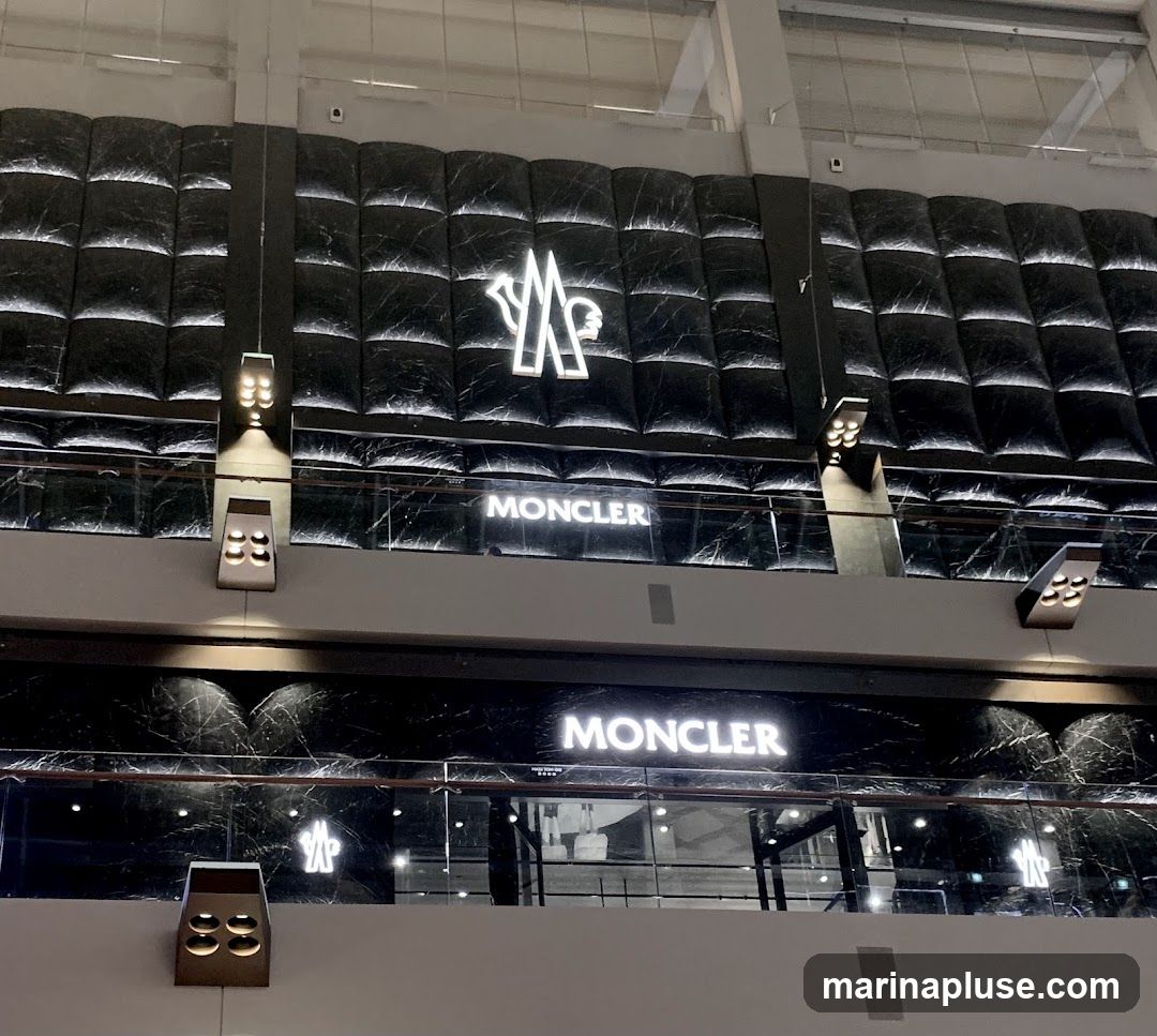 The image shows the exterior of a Moncler store. The store features a dark, marble-patterned facade with illuminated Moncler logos and signage. The upper level has a large, stylized Moncler logo against a quilted, dark background. Below, a prominent "MONCLER" sign is visible, along with more logos and architectural lighting. The lower level also displays "MONCLER" signage and logos, with glass storefronts revealing glimpses of the interior.