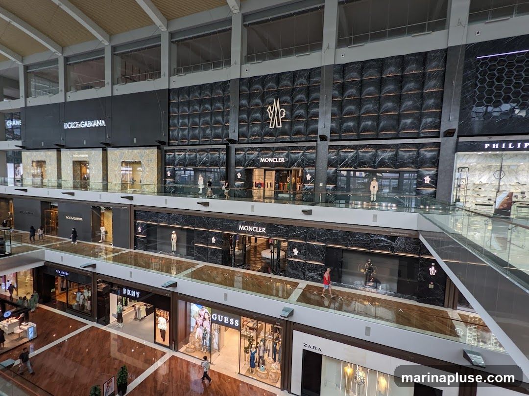 An overhead view of a multi-level luxury shopping mall. Visible storefronts include Dolce & Gabbana, Moncler, and Guess. People are seen walking on the elevated walkways between stores. The architecture features marble and dark paneling with prominent brand signage.