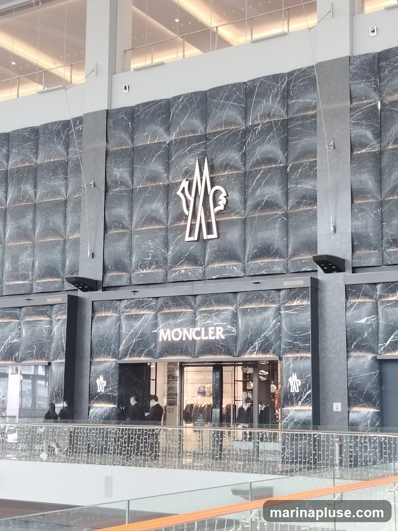 The exterior of a Moncler store with a marble-patterned facade. The Moncler logo is prominently displayed above the entrance and on the sides of the building. Through the glass entrance, racks of clothing and mannequins are visible. Several people are standing near the entrance.