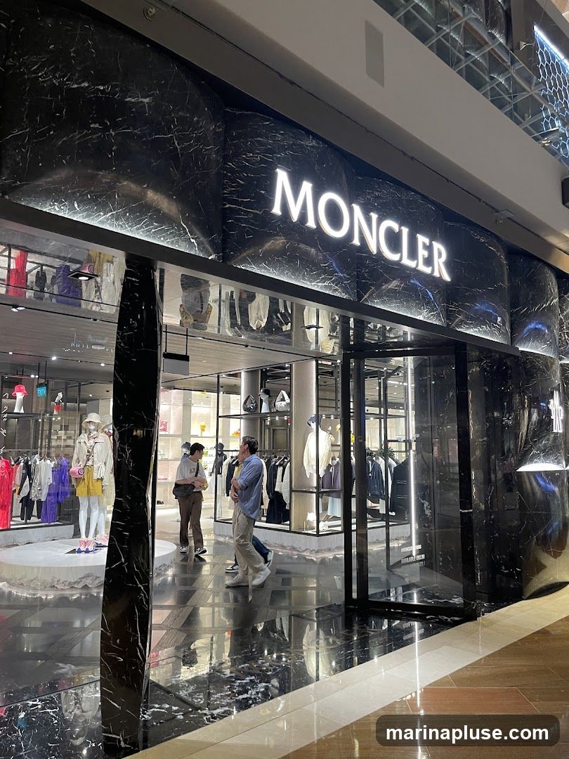 The exterior of a Moncler store with the brand name prominently displayed in illuminated white letters on a black marble facade. The entrance is visible, with mannequins displaying clothing inside. Several people are seen entering and browsing the store.