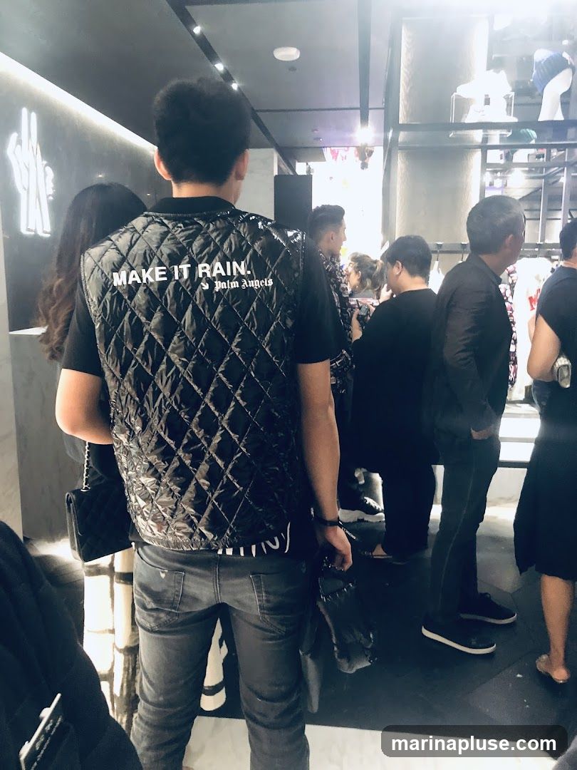A group of people are standing inside a store. The person in the foreground is wearing a black quilted vest with the text "MAKE IT RAIN." and "Palm Angels" printed on the back. Other people are visible in the background, browsing merchandise.