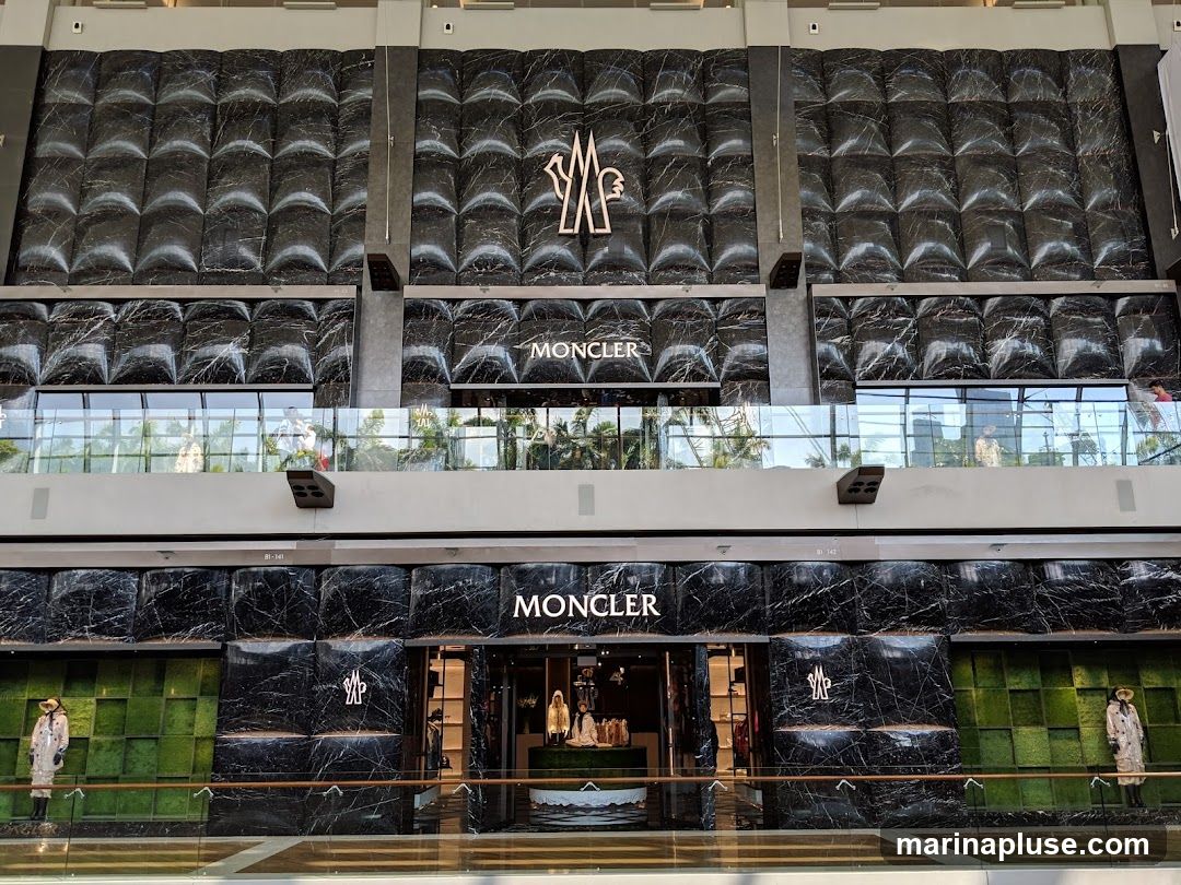 The exterior of a Moncler store with a black marble facade. The Moncler logo and name are prominently displayed above the entrance. Mannequins dressed in Moncler clothing are visible in the display windows. The store is located in a modern indoor shopping mall.