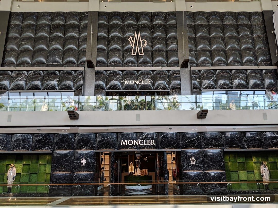 The exterior of a Moncler store with a black marble facade. The Moncler logo and name are prominently displayed above the entrance. Mannequins dressed in Moncler clothing are visible in the display windows. The store is located in a modern indoor shopping mall.