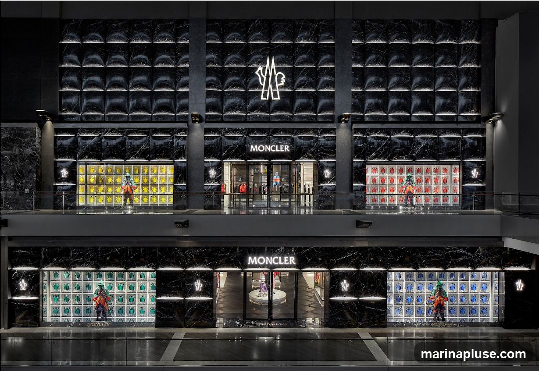 A wide shot of a Moncler store with two levels. The facade is black marble with illuminated Moncler logos and signage. Each level features a central entrance flanked by display windows. The upper level windows showcase yellow and red jackets, while the lower level windows display green and blue jackets. Mannequins are placed in front of the displays.
