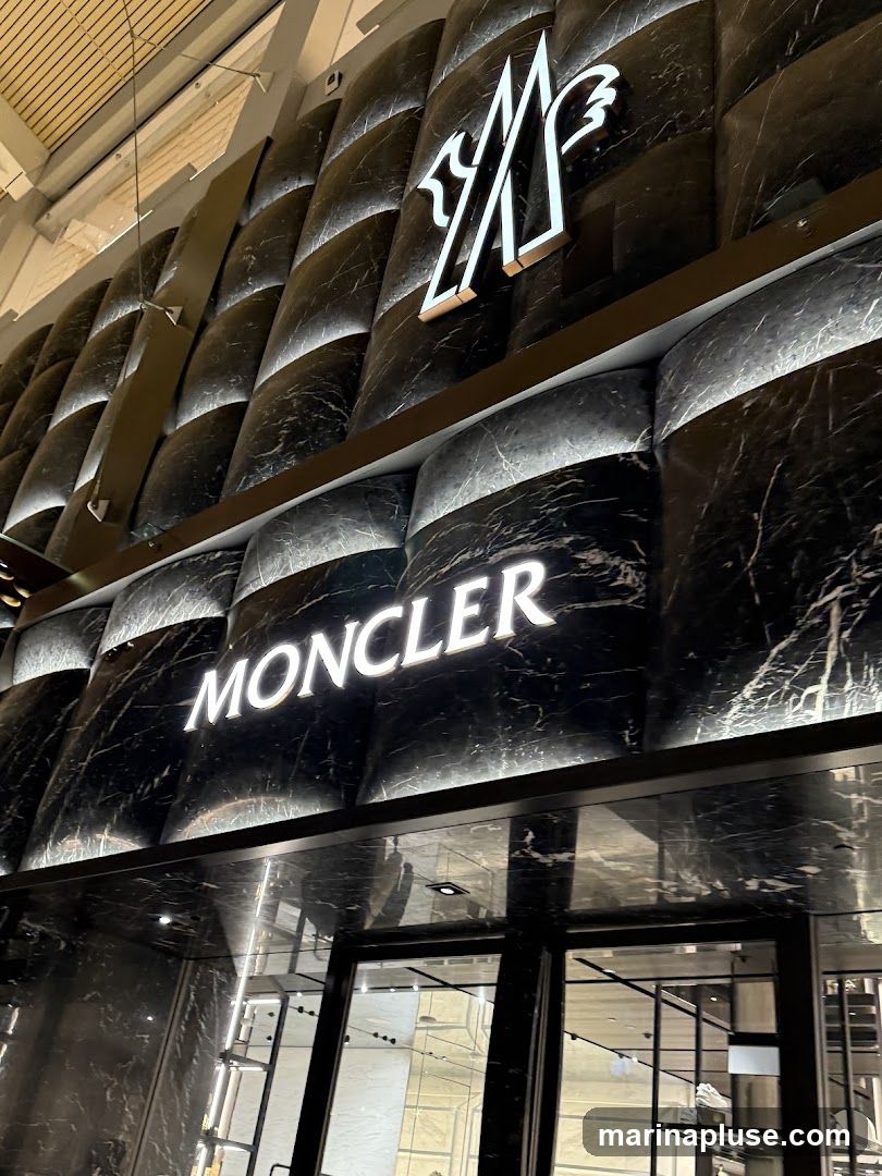The exterior of a Moncler store features a dark, marble-patterned facade with illuminated signage. The Moncler logo and name are prominently displayed in white, backlit letters. The entrance appears to be a glass door, reflecting the interior of the store. The architectural design of the facade is textured and modern, with curved panels and integrated lighting.