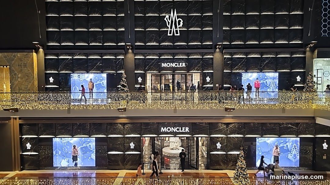 A wide shot of a Moncler store with two levels visible. The upper level features a large Moncler logo above the entrance, flanked by digital screens displaying models in winter clothing. The lower level also has a Moncler sign above its entrance, with digital screens on either side showing similar winter-themed displays. Both levels are decorated with festive string lights. People are seen walking around and inside the store.