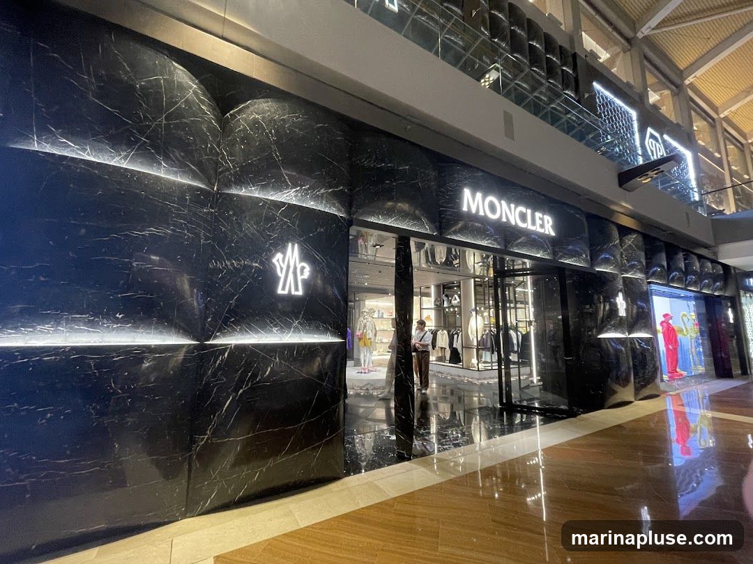 The exterior of a Moncler store with a black marble facade. The Moncler logo is illuminated in white above the entrance. Inside, clothing racks and mannequins are visible. The store is located within a modern indoor shopping mall.