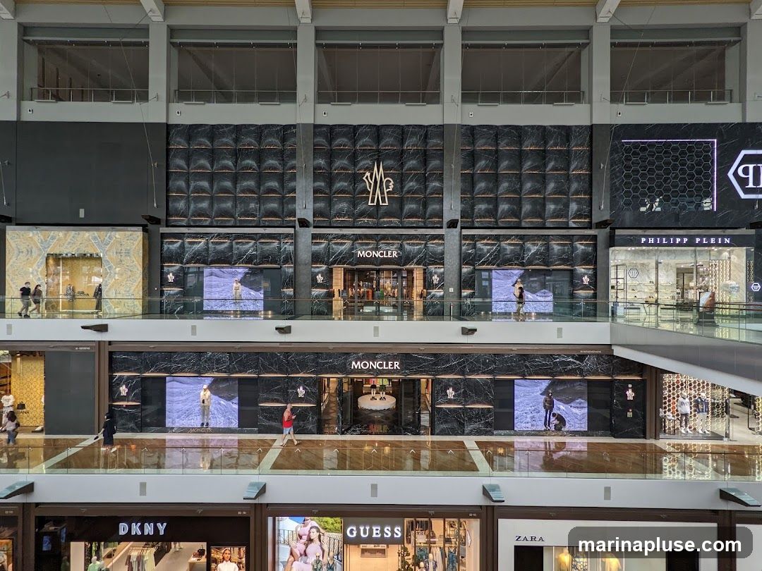 A multi-level interior view of a modern shopping mall featuring several high-end fashion brands. The image showcases the facades of Moncler, Philipp Plein, DKNY, Guess, and Zara stores. Moncler stores are prominently displayed with their logo and marble-patterned walls, featuring digital screens with models. Other stores have their distinct signage and window displays visible. People are seen browsing and walking on the different levels of the mall.