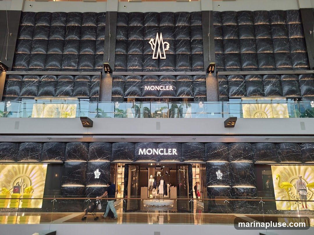 The exterior of a Moncler store, featuring a dark, marble-patterned facade with the Moncler logo prominently displayed. The entrance is visible, with mannequins and merchandise displayed inside. Large illuminated panels with the Moncler logo are on either side of the entrance.