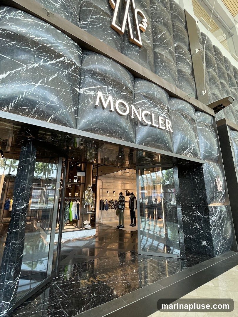 The exterior of a Moncler store with a marble-patterned facade. The Moncler logo and name are prominently displayed on the building. Through the glass entrance, clothing and mannequins are visible inside the store.