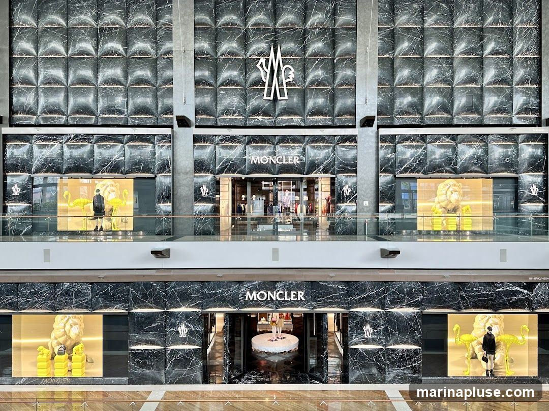 A multi-level store facade with a marble-patterned exterior. The Moncler logo is prominently displayed on the upper and lower levels. The storefront features large display windows showcasing mannequins and artistic installations, including yellow flamingos and a lion sculpture.