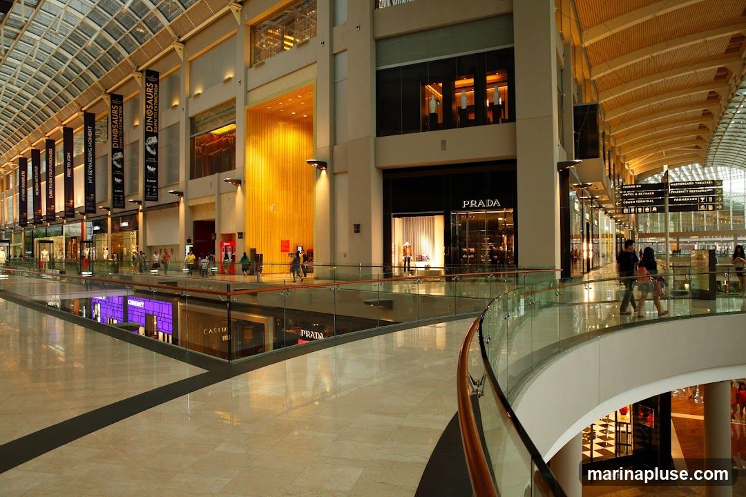 A wide shot of the interior of a large, modern shopping mall. The image shows multiple levels with shops, escalators, and a glass railing overlooking a lower level. The ceiling is a glass and metal structure, allowing natural light to filter in. Banners hang from the ceiling, advertising various events and promotions. A prominent Prada store is visible, as well as signs for other establishments like a casino, hotel, and restaurants. People are seen walking around the mall.