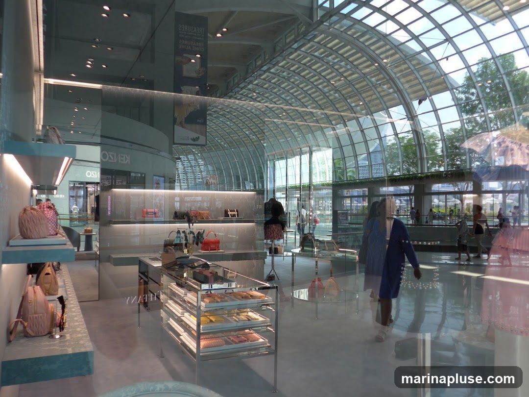A well-lit interior of a luxury store, likely a handbag boutique, with a modern and minimalist design. The store features shelves displaying various handbags and accessories. The floor-to-ceiling glass windows offer a view of a bustling atrium with a distinctive arched glass roof and people walking by. The reflection on the glass creates a layered effect, showing both the store's interior and the exterior environment.