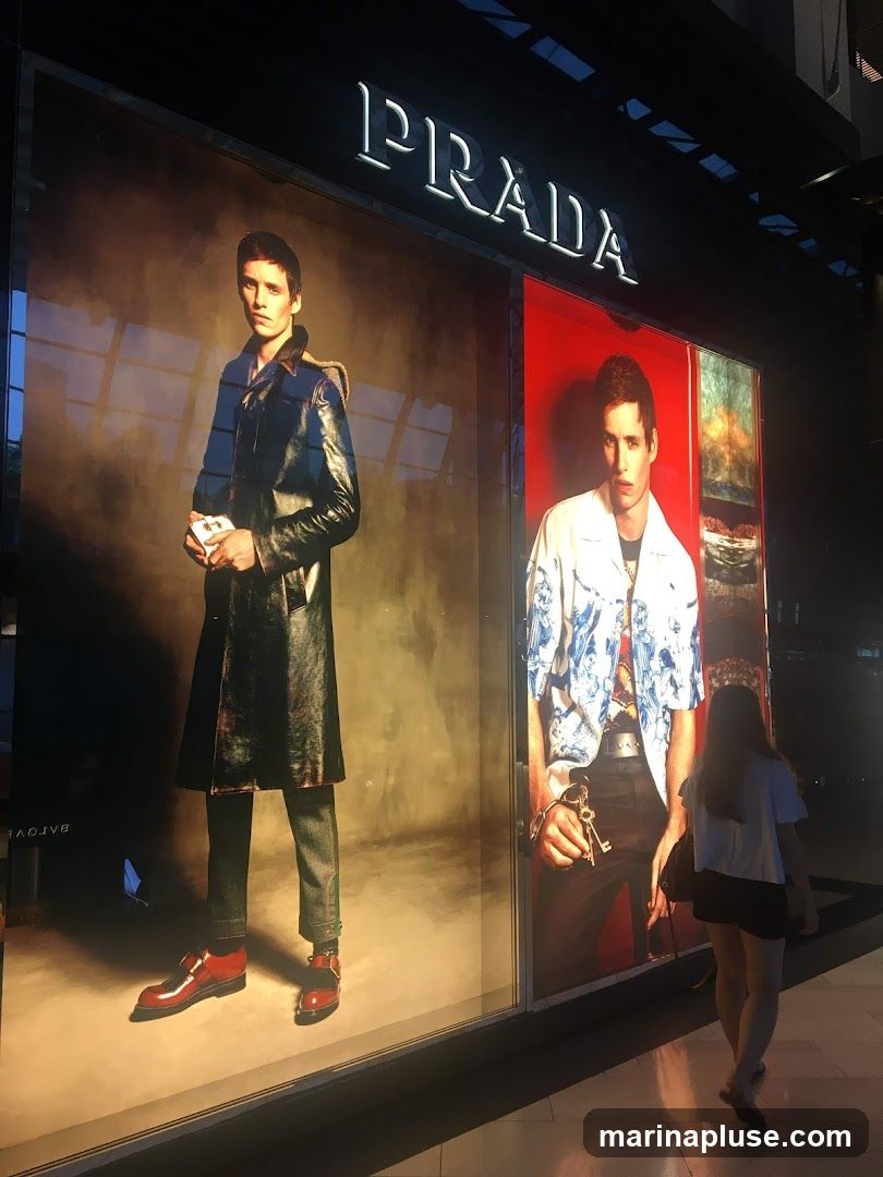 A storefront window display for Prada featuring two large advertisements of a male model. The model on the left wears a dark trench coat and red shoes, while the model on the right wears a patterned shirt and holds keys. The Prada logo is visible above the display. A person walks past the window.