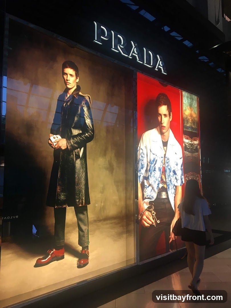 A storefront window display for Prada featuring two large advertisements of a male model. The model on the left wears a dark trench coat and red shoes, while the model on the right wears a patterned shirt and holds keys. The Prada logo is visible above the display. A person walks past the window.