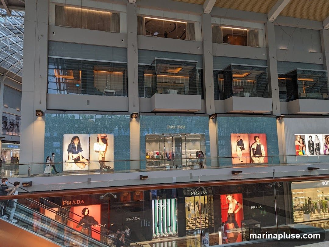 A multi-level interior view of a shopping mall with several luxury brand stores visible. On the upper level, Miu Miu has a storefront with displays and mannequins. Below, Prada and Bottega Veneta storefronts are prominent, featuring large advertisements with models and product displays. An escalator is visible in the foreground.
