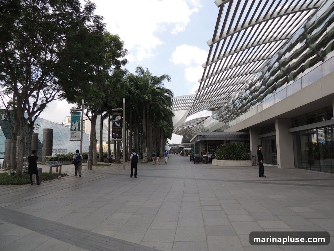 A wide paved walkway leads past a modern building with a distinctive, undulating roof structure. Palm trees and other foliage line the walkway, creating a tropical ambiance. Several people are visible walking along the path and near the building entrances. The architecture suggests a high-end shopping or entertainment complex.
