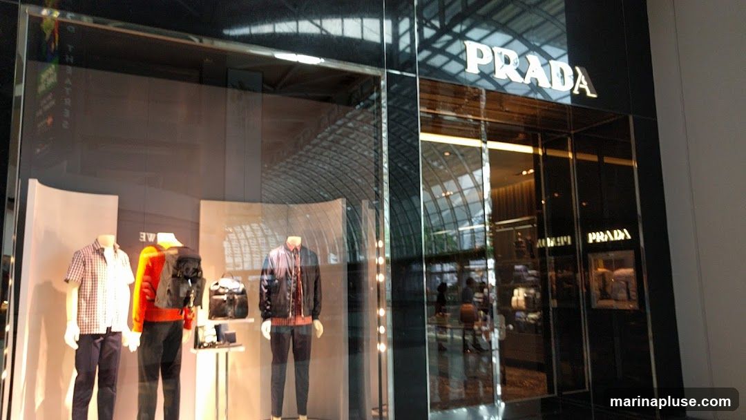 The exterior of a Prada store with mannequins displaying clothing in the window. The Prada logo is visible above the entrance.
