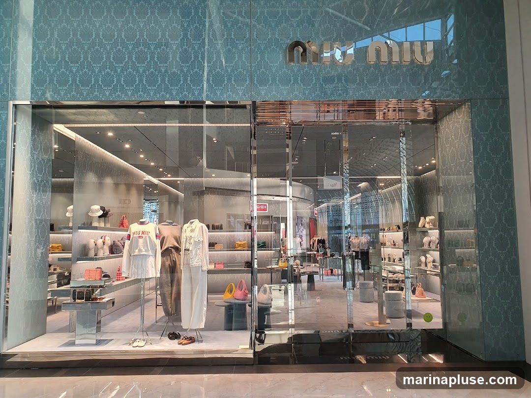 A well-lit storefront with large glass windows displaying mannequins dressed in fashionable clothing and accessories. The store's name, "MIU MIU," is prominently displayed in large, metallic letters above the entrance. The interior is visible through the glass, showing shelves stocked with various items and more displays.