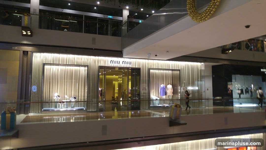The exterior of a Miu Miu store in a shopping mall. The storefront features a central entrance with glass doors, flanked by display windows. The facade is decorated with shimmering, light-colored curtains. Mannequins displaying clothing are visible in the windows. The Miu Miu logo is prominently displayed above the entrance.