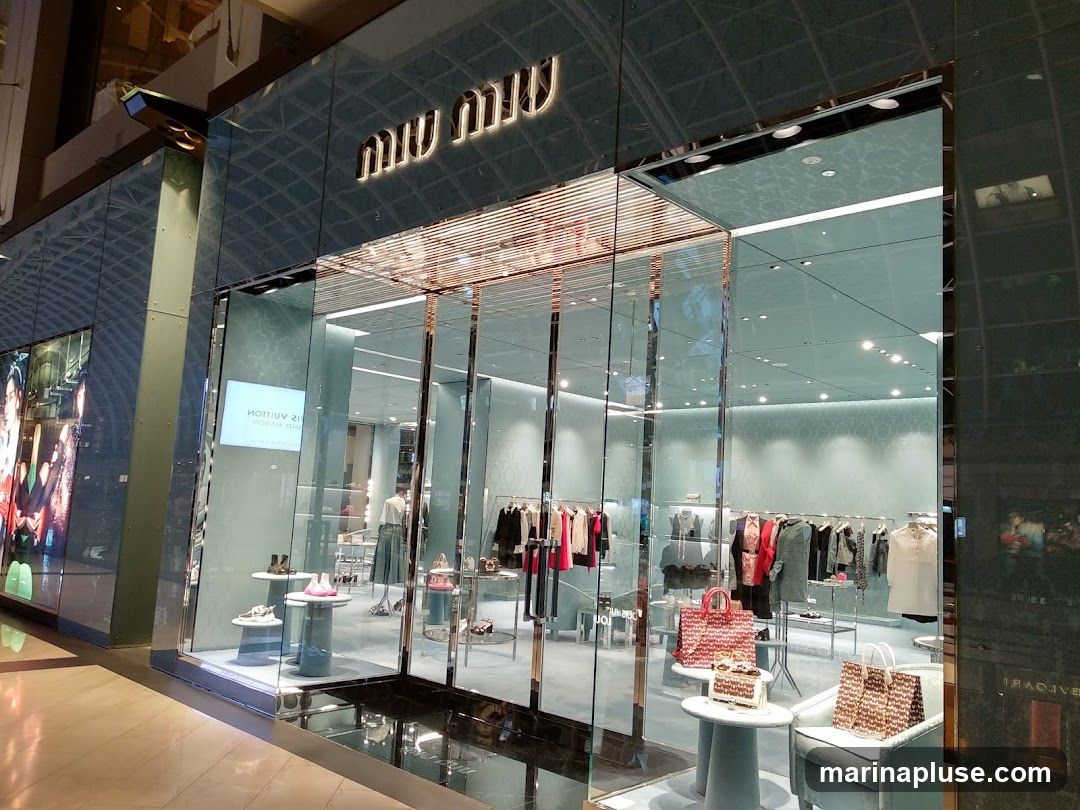The exterior of a Miu Miu store with a large glass facade. The Miu Miu logo is visible above the entrance. Inside, clothing and accessories are displayed on racks and shelves.