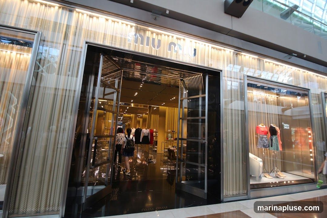 The exterior of a Miu Miu store with a large glass entrance and display windows. The Miu Miu logo is visible above the entrance. Inside, mannequins display clothing and accessories. The store features a luxurious interior with gold-colored curtains and reflective surfaces.