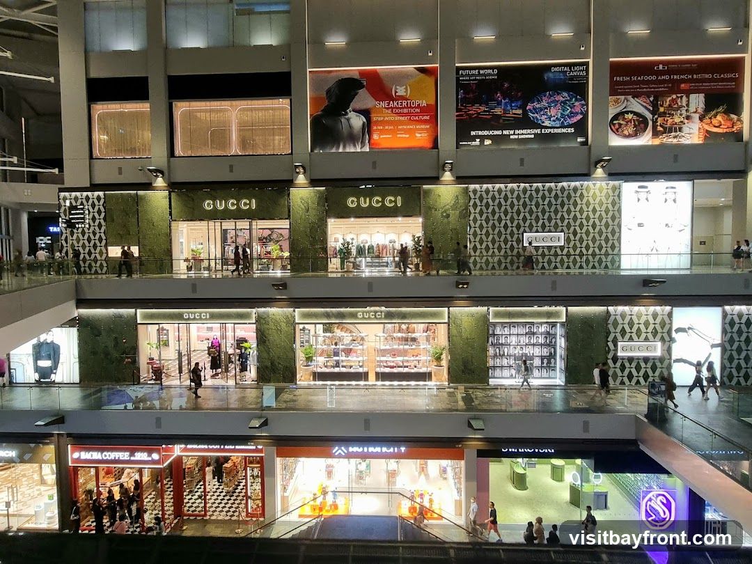 The image shows multiple levels of a modern shopping mall. Several Gucci stores are visible on the upper levels, with their distinctive green facade and signage. Above these stores, large digital billboards display advertisements for an exhibition called "SNEAKERTOPIA," an immersive experience called "FUTURE WORLD DIGITAL LIGHT CANVAS," and a restaurant offering "FRESH SEAFOOD AND FRENCH BISTRO CLASSICS." On a lower level, a store labeled "BACHA COFFEE 1810" is visible, along with other retail spaces. People are seen walking on the elevated walkways and escalators, browsing in the stores, and interacting in the mall environment.