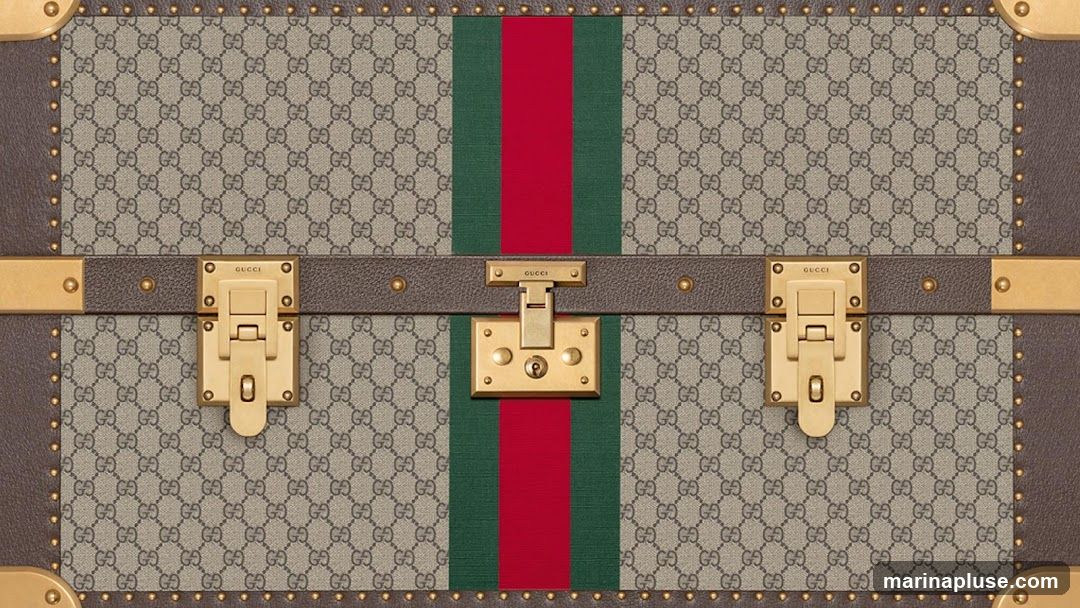 A close-up view of a Gucci trunk, featuring the brand's signature GG monogram pattern in beige and brown. A central stripe of red and green runs vertically down the trunk, accented by gold-toned hardware, including latches and a lock.