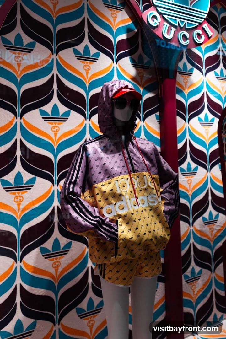 A mannequin is displayed in a store window wearing a purple and yellow Adidas x Gucci tracksuit. The jacket features the Adidas logo prominently on the front and the Gucci logo repeated throughout. The background is a colorful, retro-inspired pattern with Adidas trefoil logos and Gucci motifs. A large sign with the Adidas and Gucci logos is visible above and to the right of the mannequin.