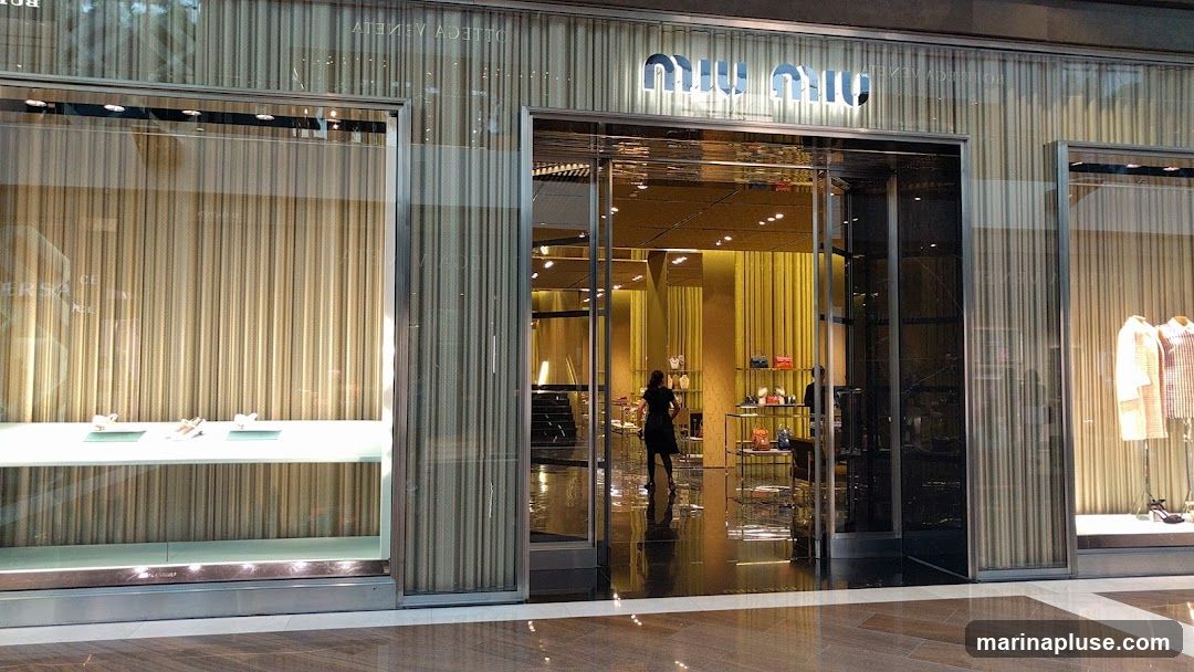 The exterior of a Miu Miu store. The storefront features a large glass entrance with a silver frame. Above the entrance, the Miu Miu logo is displayed in illuminated silver lettering. The facade is decorated with vertical, textured panels in shades of beige and gold. To the left of the entrance, a display window showcases shoes on a white shelf. To the right, another display window features clothing on mannequins. Inside the store, a woman in a black dress is visible, along with shelves displaying handbags.