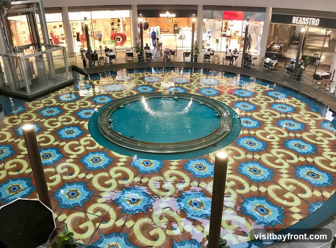 An overhead view of a circular indoor fountain with a patterned base featuring the Gucci logo. The fountain is surrounded by shops and seating areas.