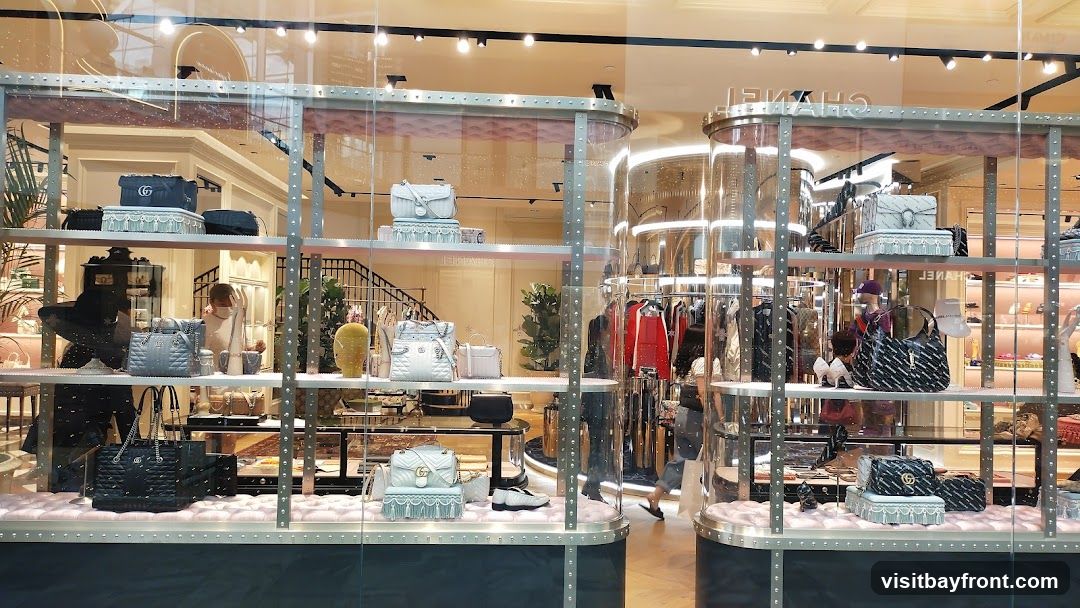 A well-lit display window showcases a variety of luxury handbags and accessories. The display features multiple shelves with handbags of different sizes, colors, and styles, including quilted leather bags and bags with prominent brand logos. Some shelves also display shoes and other small accessories. The background shows a glimpse of the store's interior, with clothing racks and a staircase. The overall presentation is elegant and sophisticated, typical of a high-end fashion boutique.