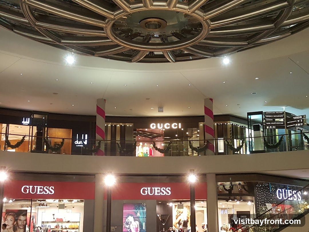 An interior view of a shopping mall with multiple storefronts visible on different levels. Prominent brands like GUCCI and GUESS are displayed. The architecture includes a large circular ceiling feature and decorative pillars.