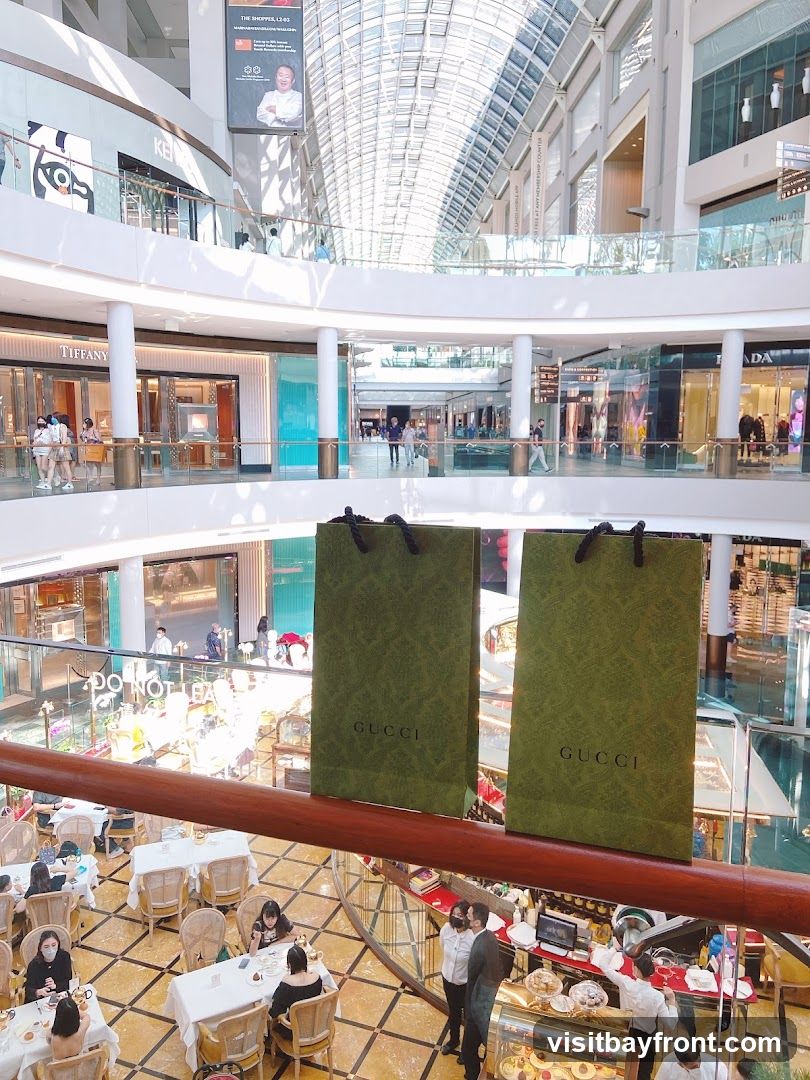 A view from above inside a multi-level shopping mall. Two green Gucci shopping bags are prominently displayed in the foreground. Below, a dining area with white-clothed tables and chairs is visible, with patrons seated. Further down, a display case with pastries and a counter area with staff are also present. The mall features a modern architectural design with a glass ceiling and multiple levels of shops, including Tiffany & Co.