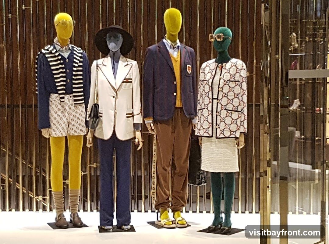 Four mannequins are displayed in a store window. The mannequins are dressed in various outfits. The first mannequin on the left wears a blue and white striped sweater over a collared shirt, white shorts with a repeating logo pattern, yellow tights, and brown loafers. The second mannequin wears a white blazer with gold trim, a blue shirt, and dark blue pants, accessorized with a black hat and a black handbag. The third mannequin is dressed in a navy blazer over a yellow sweater vest, a collared shirt, and brown pants, with yellow sandals. The fourth mannequin wears a white jacket with a repeating logo pattern, a white textured skirt, and green tights, accessorized with large sunglasses and black shoes. The background consists of vertical metallic rods.