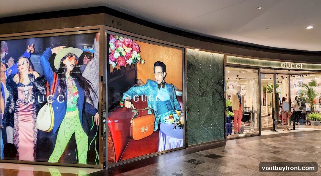 The exterior of a Gucci store with large display windows featuring fashion advertisements. The store entrance is visible with mannequins and merchandise displayed inside. The Gucci logo is prominently displayed above the entrance.