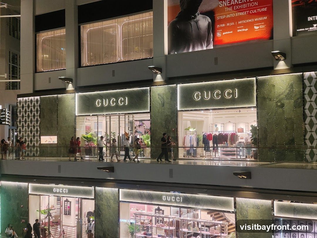 A multi-level Gucci store with prominent signage on each level. The facade features green marble-like panels and illuminated Gucci logos. People are seen walking on a balcony in front of the upper-level store, which displays clothing on racks. The lower level also shows merchandise through its windows.