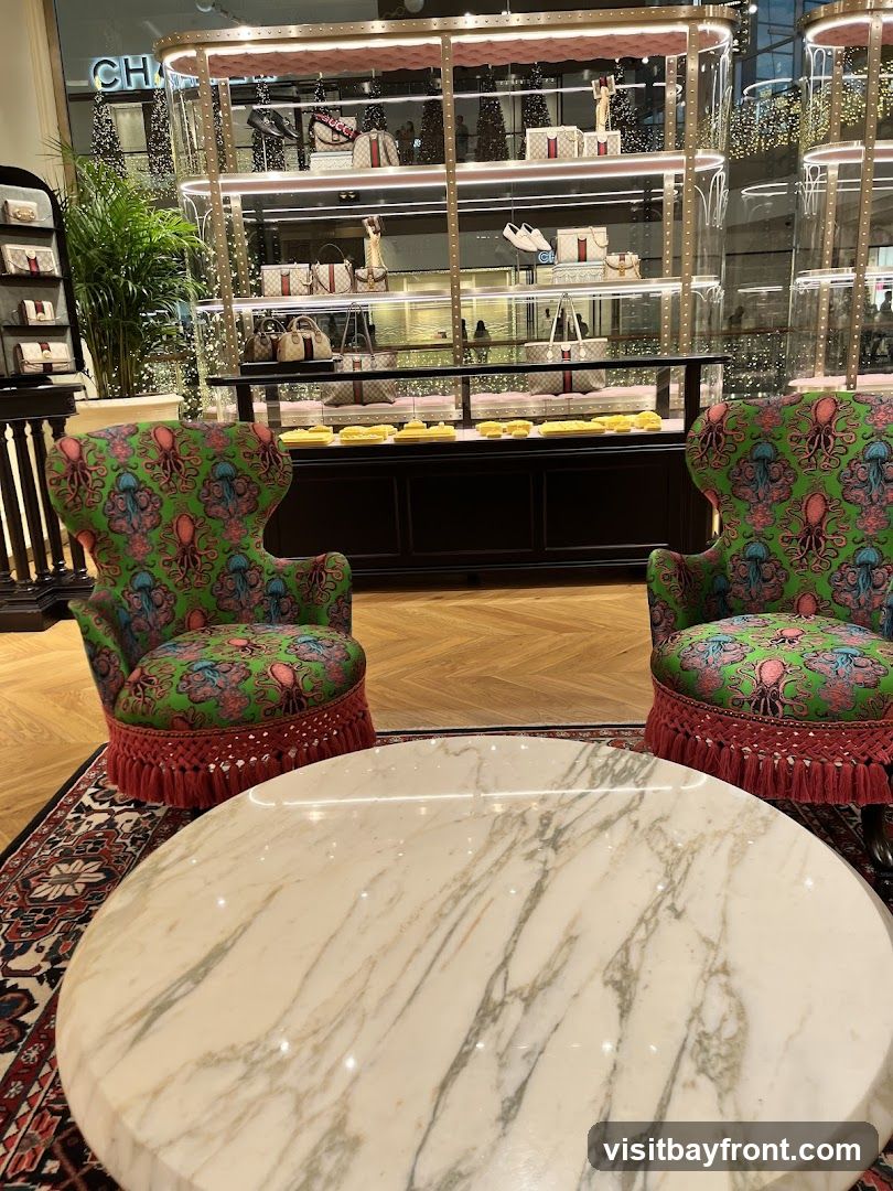 The image shows the interior of a luxury retail store. In the foreground, a round marble coffee table sits on an ornate rug. Behind the table are two ornate armchairs upholstered in a green fabric with a pink octopus-like pattern and red fringe. In the background, illuminated display cases showcase handbags, shoes, and other accessories. The store appears to be decorated for a holiday, with string lights visible.