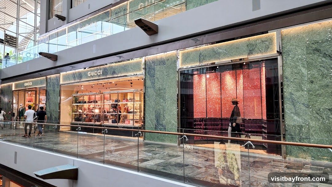 The exterior of a Gucci store with its name prominently displayed above the entrance. The store features large display windows showcasing handbags and accessories. The facade is decorated with green marble accents and illuminated signage. A person is seen walking past the store.