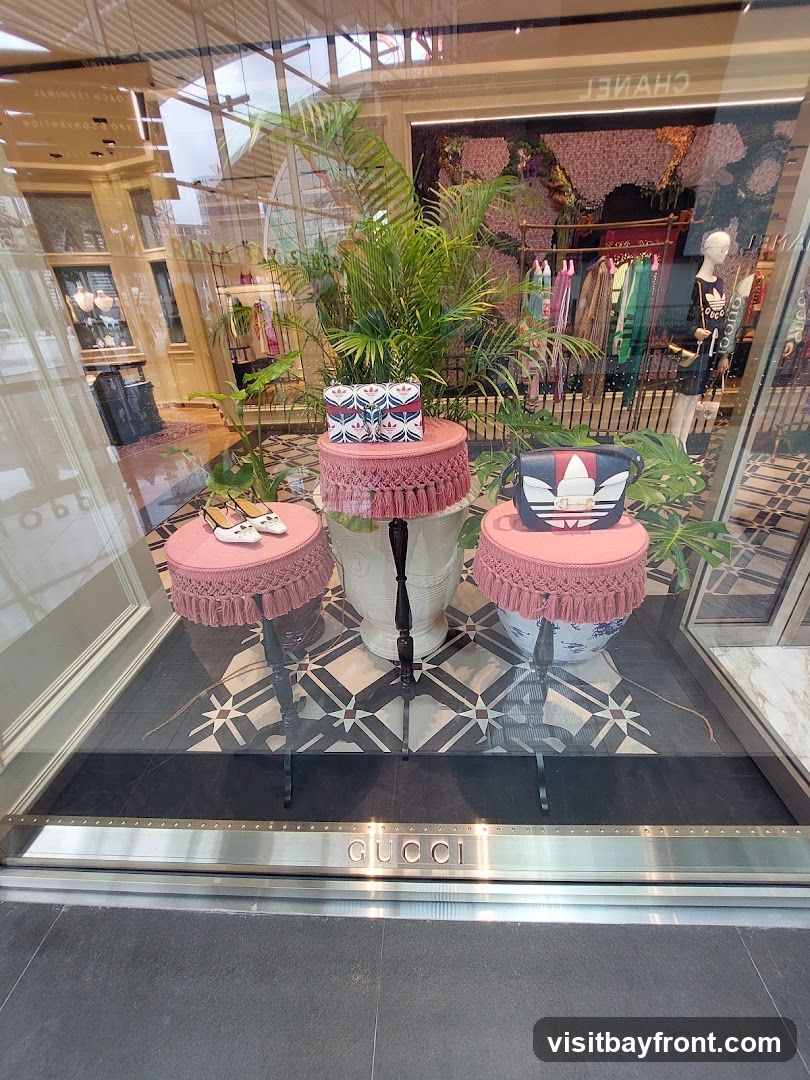 A storefront window display for Gucci, featuring handbags, shoes, and clothing. The display includes decorative plants and pink stools. The Gucci logo is visible on the exterior of the building at the bottom of the window.