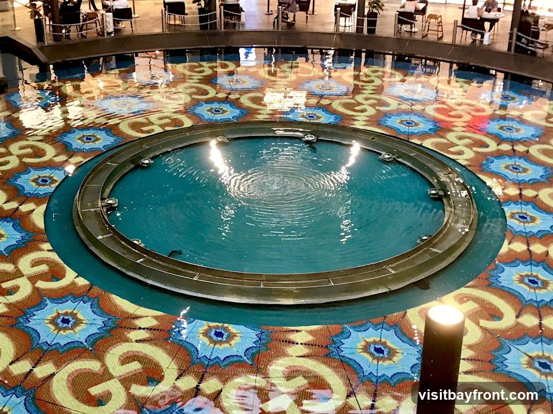 A circular fountain with teal water is centrally located on a floor with a colorful, patterned mosaic. The pattern features repeating stylized floral motifs and the interlocking letters 'GG'. Reflections of people and lights are visible on the water's surface. In the background, there are seating areas with tables and chairs, suggesting an indoor public space like a mall or hotel lobby.