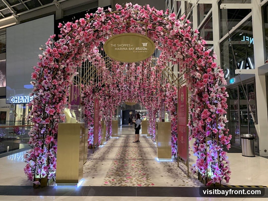 A decorative archway covered in pink cherry blossoms and hanging beads, leading down a walkway lined with display cases. The Shoppes at Marina Bay Sands Singapore logo is visible on an oval sign at the top of the arch. In the background, store signs for Chanel and other retailers are visible.