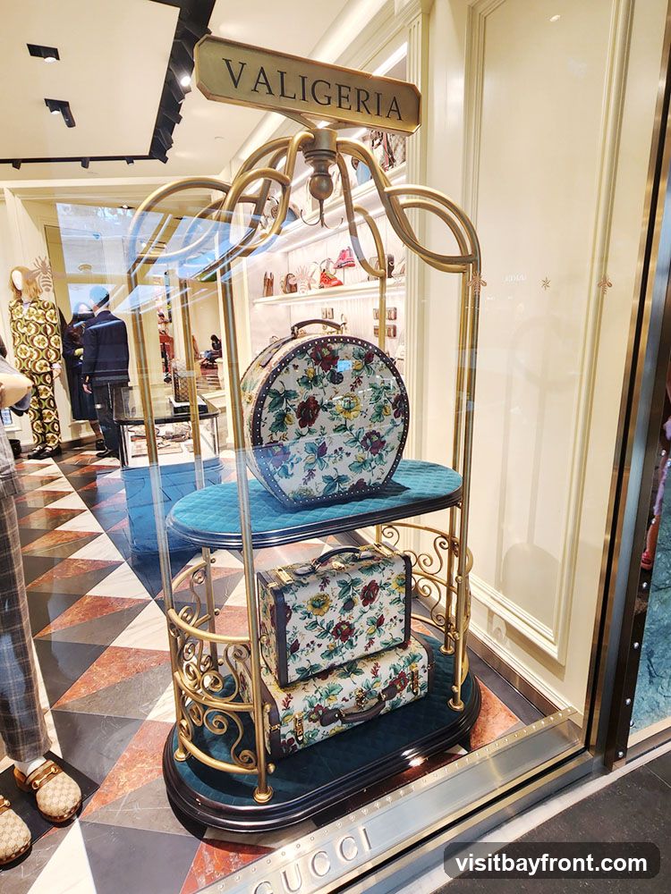 A display window featuring Gucci luggage. The display includes a round hatbox and two suitcases, all adorned with a floral pattern. The items are presented on a gold stand with teal velvet shelves. The word "VALIGERIA" is visible on a sign above the display. The Gucci logo is visible on the bottom of the display window.