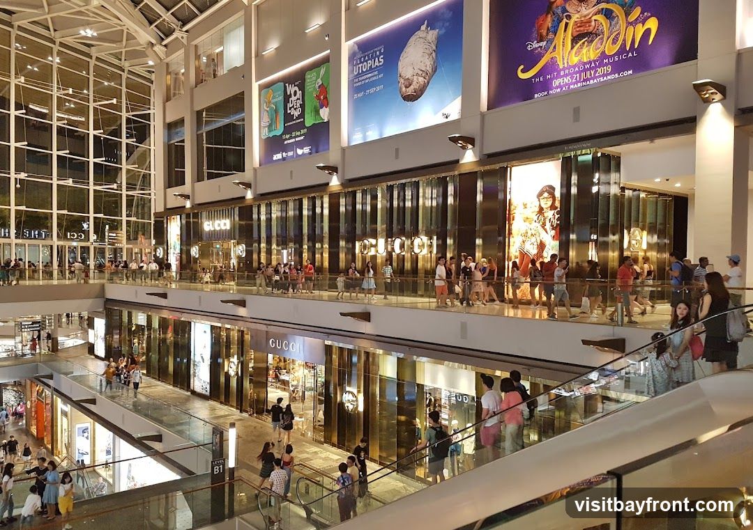 A bustling interior of a multi-level shopping mall with escalators and shoppers. Several large advertisements are displayed on the upper walls, including one for Disney's Aladdin musical and another for "Floating Utopias." Prominent Gucci storefronts are visible on multiple levels, with their logos clearly displayed.
