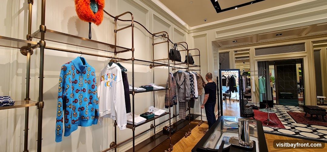 A well-lit interior of a high-end clothing store. Racks display a variety of apparel, including sweaters, hoodies, jackets, and pants. Accessories like handbags are also visible. A man is browsing the clothing racks. The store features elegant decor with patterned rugs and polished wooden floors.